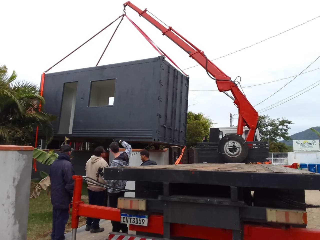 Munck içando um módulo de casa container preto enquanto a equipe orienta a descida
