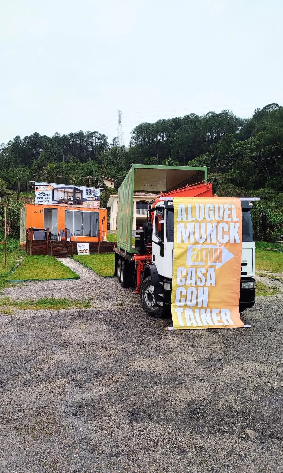 Caminhão Munck com a faixa “Aluguel Munck aqui” ao lado de uma casa container laranja